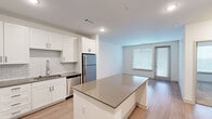 This well-designed one-bedroom, one-bathroom residence offers a thoughtfully organized single-floor layout that maximizes functionality and comfort. The open-concept living and kitchen area serves as the heart of the home, featuring wood-look plank flooring that extends throughout the main space. The kitchen is outfitted with crisp white shaker-style cabinetry, a classic subway tile backsplash, dark quartz countertops, and a full suite of stainless steel appliances including a refrigerator, range, microwave, and dishwasher. A central island with matching quartz countertop provides additional prep space and casual seating. The living area flows naturally toward a private entry door flanked by windows fitted with horizontal blinds, allowing ample natural light to brighten the space. The bedroom is carpeted for comfort, features a ceiling fan with a window overlooking greenery, and connects directly to the bathroom and a dedicated walk-in closet. The bathroom includes a tub-shower combination with white tile surround, and the adjacent closet offers built-in shelving for organized storage. Recessed lighting throughout the main areas and clean white walls contribute to a bright, modern aesthetic that makes efficient use of the available space.