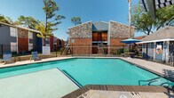 Located at 1800 FM 1092 Rd in Missouri City, Texas, this single-level outdoor amenity space centers around a generously sized swimming pool that serves as the focal point of the complex's communal area. The pool is framed by a tiled stone deck, offering ample space for lounging and relaxation under the Texas sun.

A standout feature of the area is the pergola-covered outdoor lounge, which provides welcome shade and houses a tiled bar counter with high-top seating — an ideal setting for casual gatherings. Adjacent to the pergola is a covered gazebo area with additional seating, umbrellas, and tables arranged for comfort and convenience.

The surrounding architecture blends brick and stone facades with board-and-batten siding accents, giving the complex a classic yet well-maintained character. Mature trees — including tall pines and palms — line the perimeter, adding natural shade and a lush backdrop to the outdoor environment. The entire pool area is enclosed by a wrought iron safety fence, ensuring both security and an unobstructed view of the landscaped grounds.

Lounge chairs are positioned poolside for sunbathing, and the facility includes a poolside restroom or utility building for added convenience. The combination of functional amenities and open-air design makes this a well-equipped outdoor recreation space suited for warm-weather enjoyment year-round.