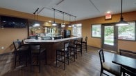 Located at 3594 N Cederblom St in Coeur d'Alene, Idaho, this single-floor commercial dining and lounge space offers a warm, inviting atmosphere designed for comfortable gathering and dining. The interior is finished with rich wood-look flooring throughout and painted in earthy golden-amber tones that lend a cozy, approachable character to every area.

The main dining room features a generous arrangement of dark wood tables paired with ladder-back chairs, providing flexible seating for groups of varying sizes. Along one wall, a continuous upholstered booth — upholstered in a classic striped fabric — runs the length of the room, offering comfortable banquette-style seating beneath a row of framed artwork and pendant track lighting.

A curved bar counter anchors one side of the space, fitted with bar-height seating and illuminated by pendant lights hanging from a track rail above. The bar opens to a visible service and kitchen area behind it, with windows nearby that bring in natural light and views of the greenery outside.

A dedicated lounge nook provides a more relaxed setting, furnished with a sofa, ottomans, and a floor lamp, accented by large vintage-style beverage posters that add a retro, social ambiance. A wall-mounted television and a high-top table complete this casual corner. Track lighting throughout the entire space ensures even, adjustable illumination across all seating areas.