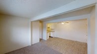 This single-floor space offers an open and functional layout that connects the main living area directly to the kitchen, creating a cohesive flow throughout. The living area is finished with neutral-toned carpet, providing a comfortable and understated foundation for the room. A textured popcorn ceiling runs throughout, complementing the clean, light-colored walls that contribute to a bright and airy atmosphere.

The transition into the kitchen area is defined by a subtle change in flooring, shifting from carpet to smooth vinyl that mimics a wood-like appearance. The kitchen is equipped with white cabinetry offering ample storage, a countertop workspace, and a sink positioned along the wall. White appliances are integrated seamlessly into the cabinetry, maintaining a consistent and tidy aesthetic. A ceiling fan with a light fixture is installed above this area, providing both air circulation and overhead illumination.

A doorway adjacent to the living area suggests access to additional rooms, such as a bedroom or hallway. Overall, the space presents a straightforward, practical design with a neutral palette that offers a clean and move-in-ready environment.