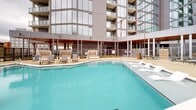 Situated in the heart of downtown Atlanta, this generously sized rooftop amenity deck offers a premier outdoor experience with sweeping views of the city skyline. The centerpiece of the space is a large, geometrically shaped swimming pool with crystal-clear turquoise water, in-pool lounge chairs, and easy-access entry steps — designed for both relaxation and recreation. The pool deck is finished in neutral-toned concrete pavers that extend across the expansive surrounding terrace, accommodating rows of modern chaise lounges and shaded umbrella seating areas ideal for sunbathing and socializing. A dedicated lounge zone features comfortable outdoor sofas and canopied daybeds, providing a more intimate setting within the open-air environment. String lights and tall pole lighting fixtures are thoughtfully placed throughout, extending the usability of the space into the evening hours. The deck wraps around the adjacent residential tower, whose glass-and-panel facade rises prominently above, offering architectural context and partial shelter. Perimeter railings frame unobstructed panoramic views of the Atlanta skyline, neighboring high-rises, and active urban development. This rooftop retreat seamlessly blends resort-style amenities with an energetic downtown backdrop.