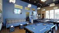 This contemporary single-floor recreational space is thoughtfully designed for socializing and entertainment. The open-concept layout is anchored by a full-size billiard table at its center, complemented by a ping-pong table nearby, making it an ideal destination for friendly competition. Bold cobalt-blue accent walls contrast striking against a dark ceiling, lending the space a modern, upscale atmosphere. Dramatic pendant lighting — including cylindrical and rectangular fixtures — hangs at varying heights, creating layers of ambient illumination throughout. A comfortable lounge area features a patterned sofa flanked by geometric accent stools and faces a wall-mounted flat-screen television, offering a relaxed setting for viewing and conversation. A second mounted screen is visible above a sleek shelving unit adorned with decorative objects, adding visual interest to the blue feature wall. Clerestory-style windows placed high on the walls allow additional natural light to filter in without compromising privacy. A wet bar or kitchenette area is visible in the background, enhancing the space's functionality as a gathering hub. The tile flooring ties the design together with a clean, contemporary finish, completing this well-appointed amenity space.
