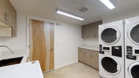 This dedicated laundry room is designed for efficiency and practicality, offering a clean, modern environment for laundry needs. The space is outfitted with multiple commercial-grade front-loading washing machines and stackable washer-dryer units, providing substantial capacity to accommodate high usage. A built-in utility sink with a sleek faucet is integrated into the countertop workspace, which is finished in a bright white surface ideal for sorting and folding. Cabinetry runs both above and below the countertop, featuring a warm wood-grain laminate finish with bar-style pulls that add a contemporary touch. The upper cabinets offer generous overhead storage, while the lower cabinets provide additional organizational space. Walls are finished in a clean white, complementing the light-toned tile flooring that gives the room a bright, hygienic feel. Overhead fluorescent strip lighting ensures the space is well-illuminated throughout. A natural wood-grain door with hardware provides access to the room. The overall layout is thoughtfully organized, maximizing functional floor space while maintaining an uncluttered and orderly atmosphere well-suited for a shared or communal laundry facility.