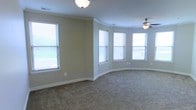 This single-floor space presents a well-designed interior highlighted by a distinctive curved bay window arrangement that floods the room with abundant natural light. The bay windows, fitted with horizontal blinds, offer flexibility in light control while providing pleasant outdoor views. The walls are finished in a warm neutral beige tone, complemented by clean white crown molding and matching baseboards that add a polished, classic touch throughout. Underfoot, plush wall-to-wall carpeting in a complementary neutral shade contributes to a comfortable and cohesive aesthetic. The ceiling is equipped with a practical ceiling fan with an integrated light fixture, promoting air circulation and supplemental lighting. A flush-mount ceiling light provides additional illumination. The curved wall design adds architectural interest and a sense of spaciousness to the room, making it feel open and inviting. Multiple windows positioned along both the straight and curved walls ensure consistent natural light from various angles, enhancing the overall brightness and airy ambiance of the space.