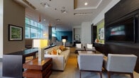 This single-floor community lounge space in Atlanta, GA (30326) presents a sophisticated, contemporary interior designed to accommodate a variety of social and leisure activities. The layout is expansive and open, seamlessly connecting several distinct functional zones within one cohesive environment.

The main entry area is characterized by floor-to-ceiling glass walls and doors that flood the space with abundant natural light while offering views of the surrounding greenery and streetscape. This transparency creates a welcoming, airy first impression. Pendant lighting in warm amber tones hangs throughout, complementing recessed ceiling fixtures and adding visual interest to the tray ceiling details above.

The central lounge area features multiple seating arrangements, including plush sofas and upholstered armchairs in neutral tones accented with colorful throw pillows. Rich wood-grain side tables and media consoles anchor the space. A dedicated media wall with a large flat-screen television and dark cabinetry provides a focal point in one section, set against a bold teal accent wall.

A separate dining and bar area includes a sleek dark table with counter-height seating, suitable for casual meetings or dining. A quieter alcove features high-backed booth seating flanked by armchairs, ideal for private conversation. Throughout, warm hardwood-style flooring and neutral carpeting unify the varied zones into a polished, inviting atmosphere.