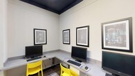 This single-floor amenity space is thoughtfully designed to serve multiple functions, seamlessly blending a business center, social lounge, and game room into one cohesive environment. The interior is defined by a striking color palette, with crisp white walls contrasting against dark-painted ceilings that lend a bold, contemporary character throughout.

The business center occupies a dedicated nook with wrap-around built-in desktops accommodating multiple workstations, each equipped with computers, keyboards, and a printer. Vivid yellow chairs provide energetic accents against the neutral tones, while framed patent-style artwork adorns the walls, adding a creative, intellectual touch. Crown molding and a high dark ceiling enhance the sense of height and sophistication in this area.

Transitioning into the main lounge, light wood-look flooring flows continuously underfoot, creating warmth and cohesion. A textured dark ceiling with recessed and track lighting sets an inviting ambiance. Comfortable leather benches with colorful throw pillows, lounge chairs, and a bean bag seat are arranged around wall-mounted flat-screen televisions, perfect for relaxation or casual gatherings. A vibrant area rug anchors the seating zone.

The game area features a foosball table positioned near large windows that invite natural light, completing this dynamic, well-appointed communal space.