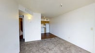 This single-floor space features an open-concept layout that seamlessly connects the living area to the kitchen. The living area is finished with neutral-toned carpet, providing a comfortable and understated foundation for the room. A textured ceiling adds subtle visual interest overhead, while wall-mounted lighting enhances the warmth of the space.

A partial dividing wall separates the living zone from the kitchen area, which transitions to durable hardwood-style flooring. The kitchen is equipped with warm-toned wooden cabinetry, white appliances including a refrigerator and stove, and ample counter space, creating a functional and practical cooking environment. A ceiling fan positioned above the kitchen area provides both ventilation and lighting.

The open flow between the two areas contributes to a sense of spaciousness, while the neutral color palette of white walls throughout creates a clean and versatile backdrop. The layout is straightforward and efficient, making it easy to navigate between functional zones. Overall, the space maintains a simple, practical aesthetic suited to everyday living.
