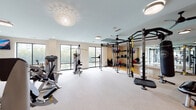 The Ezzo Kirby Med Center fitness center is a thoughtfully designed, single-floor facility that offers a comprehensive workout experience. Guests are welcomed through a polished entry lobby featuring a built-in shelving unit stocked with neatly organized amenity baskets, a beverage cooler, and a branded floor mat — setting a professional and hospitable tone from the moment of arrival.

The main fitness floor is generously proportioned and divided into distinct training zones. The cardio area is lined with large floor-to-ceiling windows that invite abundant natural light and provide calming outdoor views of surrounding greenery. A row of modern elliptical trainers, a stair climber, and a treadmill face wall-mounted flat-screen televisions, while ceiling fans overhead ensure comfortable airflow throughout workouts.

The strength and functional training zone is equally well-appointed, featuring a multi-station cable and power rack system, a heavy punching bag, TRX suspension trainers, medicine balls, and a selection of free weights on dedicated racks. Full-length mirrors along one wall enhance spatial awareness and allow users to monitor their form. A decorative pendant light fixture adds an elevated aesthetic touch to the space.

Additional amenities include a dual water fountain station, a medical-grade scale, and convenient restroom access — rounding out a fitness facility designed to meet the needs of a wide range of users.