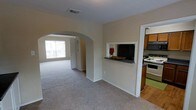 This single-story space showcases a thoughtfully designed open floor plan that promotes a natural flow between living areas. A prominent arched doorway serves as a defining architectural detail, connecting the main living space to an adjoining room while adding a classic, elegant touch to the interior. The living area is finished with wall-to-wall carpeting in a neutral tone, complementing the warm beige walls throughout.

Natural light fills the space through a large window visible beyond the arched passage, brightening the interior and creating an inviting atmosphere. The kitchen is positioned to one side and features a pass-through window with a dark granite-style counter ledge, offering a convenient connection between the kitchen and the main living area. The kitchen itself is equipped with wood-toned cabinetry, a white range, and a built-in microwave or range hood above the stove. Hard flooring in the kitchen contrasts with the soft carpeting in the adjoining rooms.

White trim and baseboards frame the neutral walls throughout, lending a clean, cohesive finish to the overall space. The layout and design details combine to create a functional and comfortable environment.