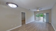 This single-floor residence is designed with a clean, contemporary aesthetic, featuring warm-toned hardwood-style flooring and neutral gray walls that create a cohesive, move-in-ready atmosphere throughout. The open-concept living and dining area is bright and spacious, equipped with a ceiling fan with integrated lighting and a large sliding glass door that opens to a private balcony, allowing natural light and greenery views to flow into the space. A pendant-lit kitchen island with a single-basin sink and light countertops anchors the kitchen, which features gray shaker-style cabinetry, white subway tile backsplash, and a full suite of stainless steel appliances including a refrigerator, range, and microwave. The layout connects seamlessly to the living area through an open pass-through arrangement. The bedroom is generously proportioned with its own ceiling fan, a window with blinds that frames outdoor greenery, and ample wall space. The bathroom presents a long, elongated layout with a dual-sink gray vanity topped with a white countertop, large mirror, tile flooring, and a full tub-shower combination, all finished in crisp white.