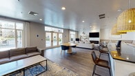 Located at 1001 Madison St in Hoboken, NJ, this single-floor amenity lounge is designed with a contemporary aesthetic that balances function and comfort. The space is unified by warm hardwood-style flooring, recessed lighting, and a neutral color palette of soft grays and whites throughout.

The main lounge area is generously proportioned, offering multiple distinct seating zones. A standout feature is the white stacked-stone fireplace wall, which anchors the room and supports a large flat-screen television above a linear electric firebox. Built-in bench seating and tulip-style accent tables complement this focal point. Nearby, a circular wood dining table with modern chairs provides a casual gathering spot, while a sectional sofa and additional leather accent chairs create a relaxed lounge environment.

Large French doors and oversized windows flood the space with natural light and offer direct views of the surrounding neighborhood streetscape and landscaping. A secondary seating nook near the windows features a plush sectional and accent chair, ideal for quiet use.

The kitchen bar area includes dark granite countertops, white cabinetry, stainless steel appliances, and distinctive woven pendant lights overhead. Bar-height seating lines the counter. Glass double doors lead to an adjacent fitness facility, making this a well-connected, multi-functional community space.