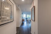 This single-floor residence showcases a thoughtfully designed modern interior with a clean, contemporary aesthetic throughout. Dark hardwood flooring runs continuously across the space, anchoring each room with warmth and visual cohesion.

The entry hallway sets an inviting tone, lined with white walls and accented by bold framed artwork. It leads seamlessly into the main living area, where large floor-to-ceiling windows and a sliding glass door open directly onto a private outdoor patio, bringing in generous natural light and views of surrounding greenery and city buildings.

The living space is characterized by an eclectic yet curated arrangement, featuring a gallery wall of framed art pieces alongside a mid-century credenza. The kitchen is outfitted with flat-front white cabinetry, a white tile backsplash, stainless steel appliances including a gas range, built-in microwave, and a French door refrigerator, all complemented by white quartz countertops.

The bedroom is a calm retreat with neutral tones, carpeted flooring, and dual windows that admit soft natural light while maintaining privacy. The bathroom presents a clean, minimalist design with dark tile flooring, a long white vanity, and a glass-enclosed shower with large-format tile surrounds. Curated artwork throughout adds personality and warmth to this polished, contemporary space.