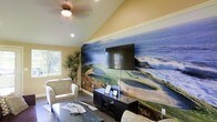 This single-floor space makes a bold visual statement with a large-scale photographic wall mural depicting a scenic coastal golf course, complete with crashing waves and lush fairways. The mural serves as the focal point of the room, framing a wall-mounted flat-screen television positioned above a dark wood media console. The vaulted ceiling adds a sense of openness and volume to the space, enhanced by recessed lighting and a ceiling fan that contributes both function and character. Warm yellow-toned walls complement the cool blues and greens of the mural, creating a lively yet relaxed ambiance. A side window allows natural light to filter in, brightening the room and offering a glimpse of surrounding greenery. Comfortable upholstered armchairs and a dark coffee table furnish the seating area, which is accented by a tall potted plant that adds a natural, organic touch. The tile flooring grounds the space with a clean, low-maintenance finish. Overall, the room blends casual comfort with a distinct visual personality, making it well-suited as a lounge or recreation area.