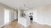 Located at 5801 Nicholson Lane in Rockville, MD, this well-appointed 2-bedroom, 2-bathroom residence offers a thoughtfully designed single-floor layout with modern finishes throughout. Light wood-look flooring extends across the main living areas, complemented by warm neutral tones on the walls and crisp white trim.

The open-concept kitchen and living area serves as the heart of the home. The kitchen features white shaker cabinetry, stone countertops, a peninsula island with an undermount sink, stainless steel appliances — including a gas range, built-in microwave, and refrigerator — and pendant lighting overhead. The adjacent living and dining areas are bathed in natural light, with large windows and a glass balcony door offering expansive outdoor views of the Rockville skyline.

The spacious primary bedroom is carpeted for comfort and includes a large picture window with scenic views. Its en-suite bathroom is finished with white cabinetry, stone countertops, and light wood-look flooring. The second bedroom is similarly carpeted and well-proportioned, served by a second full bathroom with matching white vanity and stone counters.

A hallway connects the rooms efficiently, and a dedicated closet provides additional storage. The space presents a clean, contemporary aesthetic throughout.