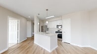 Located at 5801 Nicholson Lane in Rockville, MD, this well-appointed 2-bedroom, 2-bathroom residence offers a thoughtfully designed single-floor layout with modern finishes throughout. Light wood-look flooring extends across the main living areas, complemented by warm neutral tones on the walls and crisp white trim.

The open-concept kitchen and living area serves as the heart of the home. The kitchen features white shaker cabinetry, stone countertops, a peninsula island with an undermount sink, stainless steel appliances — including a gas range, built-in microwave, and refrigerator — and pendant lighting overhead. The adjacent living and dining areas are bathed in natural light, with large windows and a glass balcony door offering expansive outdoor views of the Rockville skyline.

The spacious primary bedroom is carpeted for comfort and includes a large picture window with scenic views. Its en-suite bathroom is finished with white cabinetry, stone countertops, and light wood-look flooring. The second bedroom is similarly carpeted and well-proportioned, served by a second full bathroom with matching white vanity and stone counters.

A hallway connects the rooms efficiently, and a dedicated closet provides additional storage. The space presents a clean, contemporary aesthetic throughout.