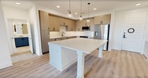 This 1,020-square-foot single-floor residence offers 2 bedrooms, 2 bathrooms, and 2 closets, delivering a thoughtfully designed modern living experience. The open-concept kitchen and living area serves as the heart of the home, showcasing light wood-grain cabinetry with matte black hardware, white quartz countertops, stainless steel appliances including a built-in microwave and dishwasher, and a large central island with pendant lighting overhead. A built-in beverage refrigerator adds a premium touch. Light wood-tone luxury vinyl plank flooring flows consistently throughout the entire unit, enhancing its cohesive, contemporary aesthetic. The primary bedroom measures 150 square feet and features a ceiling fan, direct balcony access via French doors offering open exterior views, and a dedicated walk-in closet with built-in shelving and hanging rods. The en-suite bathroom is a standout, featuring a bold blue vanity with dual sinks, matte black fixtures, an illuminated mirror, and a glass-enclosed tile shower. The second bedroom also includes a ceiling fan, a walk-in closet with built-in organization, and its own balcony access. A second full bathroom serves the additional bedroom. Recessed lighting and clean white walls complete the bright, modern ambiance throughout.