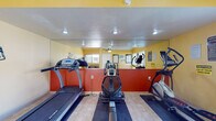 This single-floor fitness center is designed to provide a functional and motivating workout environment. The space is finished with warm yellow and bold red accent walls, a textured ceiling, and durable tile flooring throughout, lending it a vibrant and energetic atmosphere.

The main workout area is outfitted with a selection of cardio equipment, including a treadmill, a stationary rowing machine, an upright cycle, and an elliptical trainer, each positioned to allow comfortable use. A large wall-mounted mirror runs along one side of the room, visually expanding the space and allowing users to monitor their form during exercise. Motivational wall decals, including a prominent "No Pain No Gain" graphic, add an encouraging touch to the décor.

A dedicated stretching and free weights area features a floor mat and a set of dumbbells, complemented by posted stretching exercise charts for reference. Multiple large windows along the outer walls flood the space with natural light and offer views of the landscaped exterior grounds and surrounding balconies.

A small utility nook provides a built-in countertop surface, a sanitation dispenser, and additional storage, supporting the practical needs of gym users. A ceiling fan aids in air circulation, and a wall-mounted HVAC unit ensures climate control year-round. Posted safety and exercise advisory notices reflect a commitment to user well-being.