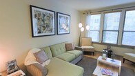 This single-floor space offers a welcoming and well-organized living environment designed with comfort and style in mind. The living area serves as the focal point of the space, featuring a light green sectional sofa adorned with neutral accent pillows, creating a relaxed yet polished atmosphere. A woven accent chair positioned near the window adds additional seating and texture to the room. Two large, boldly framed abstract artworks hang prominently on the wall, lending an artistic and contemporary character to the space. Natural light filters generously through windows fitted with slatted blinds, brightening the warm, neutral-toned walls and enhancing the overall sense of openness. A sculptural multi-globe arc floor lamp adds both ambient lighting and a modern design element to the corner of the room. The flooring features a deep-toned area rug that anchors the seating arrangement and adds warmth underfoot. A light-colored coffee table completes the living area setup, providing functional surface space. The overall ambiance is calm, curated, and inviting, with a cohesive color palette of greens, grays, and warm neutrals that tie the design elements together seamlessly.