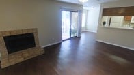 This single-floor residence offers a welcoming and functional layout designed for comfortable everyday living. The main living area is a standout feature of the home, showcasing rich, dark hardwood floors that extend throughout the open space, lending warmth and a sense of continuity. A tile-surround fireplace anchors one corner of the room, providing a classic focal point that adds both character and coziness to the space. Large sliding glass doors dominate one wall of the living area, inviting an abundance of natural light and offering a direct view to the outdoor patio or yard beyond, creating a seamless connection between indoor and outdoor living. The neutral-toned walls complement the flooring and contribute to a bright, airy atmosphere throughout. A textured ceiling adds subtle architectural detail overhead, while clean white baseboards frame the room with a crisp, finished look. An adjacent area, visible through an open pass-through or entryway, suggests additional living or dining space, further enhancing the home's open and accessible flow. Overall, the space presents a clean, move-in-ready environment that balances practical design with inviting warmth.