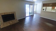 This single-floor residence offers a welcoming and functional layout designed for comfortable everyday living. The main living area is a standout feature of the home, showcasing rich, dark hardwood floors that extend throughout the open space, lending warmth and a sense of continuity. A tile-surround fireplace anchors one corner of the room, providing a classic focal point that adds both character and coziness to the space. Large sliding glass doors dominate one wall of the living area, inviting an abundance of natural light and offering a direct view to the outdoor patio or yard beyond, creating a seamless connection between indoor and outdoor living. The neutral-toned walls complement the flooring and contribute to a bright, airy atmosphere throughout. A textured ceiling adds subtle architectural detail overhead, while clean white baseboards frame the room with a crisp, finished look. An adjacent area, visible through an open pass-through or entryway, suggests additional living or dining space, further enhancing the home's open and accessible flow. Overall, the space presents a clean, move-in-ready environment that balances practical design with inviting warmth.
