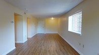 This single-floor space offers a straightforward and functional layout designed for comfortable everyday living. The main living area is defined by smooth hardwood-style flooring in a warm honey tone, lending a clean and cohesive look throughout the space. Neutral cream-colored walls provide a versatile backdrop that suits a variety of interior styles and décor choices. A large window along one wall allows natural light to fill the room, creating a bright and airy atmosphere while offering a view of the exterior brick facade. The ceiling is finished with a subtle texture and fitted with a flush-mounted light fixture, ensuring adequate illumination throughout the day and evening. White baseboards frame the perimeter of the room, adding a crisp, finished detail to the overall aesthetic. A white entry door is visible at the far end of the space, providing a clear sense of the room's depth and layout. The open floor plan and uncluttered design make this space highly adaptable, with ample room for flexible furniture arrangements. Overall, the space presents a tidy, well-maintained interior with a neutral palette that feels both practical and welcoming.