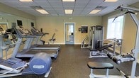 This fitness center offers a comprehensive workout environment outfitted with professional-grade equipment. The space is anchored by a lineup of Precor cardio machines, including treadmills, an elliptical, and a stationary bike, providing ample options for aerobic exercise. Complementing the cardio section, a dedicated strength training area features a multi-function cable and resistance machine along with a weight bench, catering to a full range of resistance training needs. The flooring throughout is a durable rubber composite, ideal for both equipment protection and user safety. Walls are painted in a neutral tone, contributing to a clean and focused atmosphere. The drop ceiling is fitted with recessed fluorescent lighting panels that ensure consistent, bright illumination across the entire space. A windowed door offers a natural light source and connects the room to an adjacent area. Safety is addressed with clearly posted fitness center rules and emergency contact information displayed on the wall. The overall layout is well-organized, allowing for comfortable movement between equipment stations while maintaining an efficient and functional training environment.