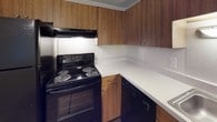 This single-floor space is designed with practicality and functionality in mind. The kitchen is equipped with dark wood-toned upper and lower cabinetry offering generous storage, a black electric coil range with overhead ventilation, a dishwasher, and light-toned countertops that extend along an L-shaped layout, providing ample workspace.

The living and dining area transitions between wood-look hard flooring and plush wall-to-wall carpeting, giving the space a sense of defined zones. A pendant light fixture illuminates the dining area, while neutral walls and a textured ceiling contribute to a clean, understated ambiance. An opening connects this area to adjacent rooms, enhancing the flow throughout the unit.

The bedrooms are carpeted and feature vertical blind-covered windows that allow natural light to filter in. One bedroom includes direct access to a walk-in closet, offering notable storage capacity.

The bathroom is finished with hardwood-style flooring, a sliding glass walk-in shower enclosure, a wide vanity countertop with an integrated sink, and a large mirror with built-in medicine cabinet. The overall palette remains neutral and consistent throughout, creating a cohesive and move-in ready environment.