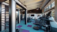 This modern fitness center is thoughtfully designed to accommodate a wide range of workout styles within a single, open floor plan. The space features soaring ceilings with exposed beam detailing and an abundance of natural light streaming through a dramatic wall of floor-to-ceiling windows, offering views of the surrounding exterior. The flooring throughout consists of teal and purple patterned rubber tiles, providing both visual energy and functional durability.

The cardio zone lines the windowed perimeter and houses a row of treadmills, elliptical machines, a stair climber, and rowing machines, each positioned to take advantage of the outdoor views. Wall-mounted flat-screen televisions provide additional entertainment options during workouts.

The strength training area features a multi-functional cable and weight machine, adjustable benches, and a well-stocked dumbbell rack. A dedicated functional training zone, defined by a Queenax rig system, includes a heavy punching bag and accessories for dynamic training, with full-length mirrors enhancing the sense of space.

A separate yoga or studio room with glass partition walls provides a quieter environment for focused training. The facility also includes a convenient amenity alcove with built-in dark wood cabinetry, a beverage cooler, a cushioned bench, open shelving, and a water fountain station, alongside accessible restroom facilities.