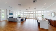 Located at 1000 Baytowne Dr in Champaign, IL, this single-floor community clubhouse offers a thoughtfully designed layout across multiple interconnected spaces. The centerpiece is a grand, light-filled great room featuring soaring windows and French doors that line an entire wall, flooding the space with natural light and providing views to the outdoors. Warm hardwood flooring flows throughout, complemented by elegant chandelier lighting and crisp white walls and trim. A grand piano anchors one corner of the main lounge, which features comfortable seating arrangements and a classic fireplace. Adjacent to the main lounge is a secondary sitting area with a large sectional sofa, a wall-mounted fireplace, and coastal-inspired artwork. A spacious bar and lounge area includes a generous white quartz island with bar seating, a wall-mounted TV, and ample room for entertaining. The service kitchen is well-equipped with stainless steel appliances, warm wood cabinetry, and beverage dispensers. A separate dining room accommodates a large farmhouse-style table beneath a modern chandelier. The fitness center is bright and airy, outfitted with strength and cardio equipment, mirrored walls, and large windows overlooking the exterior. A water station and glass-walled fitness studio complete the wellness amenities.