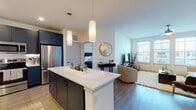 This single-floor residence at 11201 Riverbeauty Loop in Louisville, Kentucky offers a thoughtfully designed open-concept layout with contemporary finishes throughout. The kitchen is a standout feature, showcasing bold navy-blue cabinetry that extends to the ceiling, white quartz countertops, a textured geometric tile backsplash, and a full suite of stainless steel appliances. A central island with seating provides an ideal gathering space, illuminated by sleek pendant lighting overhead. Light wood-style flooring flows seamlessly through the kitchen and into the living area, where oversized windows invite an abundance of natural light and create an airy, welcoming atmosphere. The living room offers ample space for relaxing or entertaining, with ceiling fans adding to everyday comfort. The bedroom is generously proportioned, featuring plush carpeting, multiple windows, a ceiling fan, and access to a spacious walk-in closet with wire shelving for organized storage. The bathroom is finished with clean white cabinetry, quartz countertops, a large vanity mirror, and a glass-enclosed shower with tile surround. Throughout the residence, neutral tones and modern design elements combine to create a cohesive, refined aesthetic suited for comfortable everyday living.