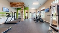 Located at 440 Cane Hill Rd in Antioch, TN, this single-floor fitness center offers a generously sized, open exercise space designed to support a comprehensive workout experience. The room features durable rubber flooring throughout, providing both safety and comfort during exercise. Large windows with decorative valances line the walls, flooding the space with natural light and offering views of the surrounding greenery, while ceiling fans and recessed lighting ensure a comfortable environment at any time of day.

The facility is well-appointed with a diverse selection of equipment, including elliptical trainers, a recumbent bike, a treadmill, and a multi-function cable machine for strength training. A power rack with a weight bench and barbell set caters to free weight exercises, while stability balls are available for functional training. Wall-mounted flat-screen TVs are strategically positioned throughout the space for entertainment during workouts.

A standout architectural feature is the rustic reclaimed wood accent columns paired with vibrant green accent walls, adding a distinctive, modern-industrial aesthetic to the space. Full-length mirrors on one wall enhance the sense of openness and allow users to monitor their form. The overall ambiance is bright, energetic, and functional, making this fitness center a well-rounded facility for residents.