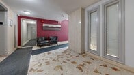 Located at 70 Park St E in Mississauga, Ontario, this building lobby serves as the main entry point for residents and visitors alike. The space is defined by its distinctive terrazzo flooring, which features a warm blend of cream and rust-toned stone fragments that lend a classic, durable character to the ground level.

Upon entry through the glass-paneled front doors, visitors are greeted by a welcoming lobby area accented by bold crimson walls that create a striking visual contrast against the lighter stone and neutral tones throughout. The seating area includes leather armchairs accompanied by contemporary cityscape artwork mounted on the feature wall, contributing to a practical yet styled atmosphere.

The lobby provides direct access to two elevators, framed by travertine-style wall panels that complement the overall aesthetic. A community bulletin board is conveniently positioned nearby, offering residents easy access to building notices and information. Textured ceilings and recessed and flush-mounted lighting fixtures ensure the space is well-illuminated throughout. The thoughtful layout efficiently guides residents from the building entrance through to elevator access, making for a functional and organized common area that reflects a well-maintained residential building in the heart of Mississauga's Port Credit neighbourhood.