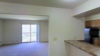 This single-floor residence offers a clean, open layout designed for comfortable everyday living. The living area is finished with plush wall-to-wall carpeting and is bathed in natural light streaming through a large sliding glass door that opens onto a private balcony — offering pleasant outdoor views of surrounding greenery. White curtain panels frame the door, enhancing the bright and airy feel of the space. The walls throughout are finished in a crisp, neutral white, creating a versatile backdrop that suits a wide range of décor styles. A wide archway connects the living area to the kitchen zone, promoting an open and connected flow between spaces. The kitchen features a raised bar-style counter with a granite-pattern surface, providing a practical breakfast bar or serving area that also acts as a natural visual divider between the kitchen and living areas. A built-in wall shelf above the bar adds additional storage or display space. The overall ambiance is light, neutral, and functional, with a layout that maximizes the sense of openness throughout the single floor. The balcony access further extends the livable space outdoors, making this a welcoming and practical home.