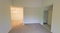 This single-floor residence presents a clean, open layout designed with simplicity and functionality in mind. The interior is finished in soft, neutral tones throughout, creating a bright and welcoming ambiance that feels both airy and adaptable to a variety of furnishing styles.

The living area is generously proportioned, featuring wall-to-wall carpeting in a warm beige hue that adds a sense of comfort underfoot. Soaring ceilings lend a notably spacious feel to the main living space, enhancing the sense of openness and volume. The transition into an adjacent room — likely a dining area — is marked by a classic chandelier, adding a refined decorative touch to the otherwise understated interior.

The space benefits from a straightforward, connected floor plan, allowing for a natural flow between areas. A tiled section is visible, suggesting a distinct functional zone such as an entryway or utility area, providing a practical contrast to the carpeted living spaces. The crisp white walls throughout serve as a blank canvas, making the space highly adaptable.

Overall, this residence offers a well-maintained, move-in-ready environment with a neutral palette, elevated ceilings, and a thoughtful layout that balances open living with defined functional zones.