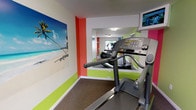 This fitness center delivers an energetic and motivating environment designed to support an active lifestyle. The space is immediately distinguished by its bold color scheme — vivid lime green, coral orange, and crisp white — applied in striking horizontal bands and accent walls that lend the room a lively, modern character.

The facility is equipped with a well-rounded selection of cardio machines, including Life Fitness treadmills and elliptical trainers, all positioned on durable, cushioned carpeted flooring. Large wall-mounted mirrors span key sections of the room, visually expanding the space and allowing users to monitor their form during workouts. Mounted flat-screen televisions are positioned for easy viewing from the equipment, providing entertainment during exercise sessions.

A large tropical beach mural adds a resort-inspired touch to one wall, creating a relaxing visual focal point. Natural light filters in through windows offering outdoor views, contributing to a bright and welcoming atmosphere. The space also includes a coat hook area with a shelf for personal belongings, a wall-mounted sanitation dispenser, and additional storage shelving — thoughtful amenities that enhance convenience. Overhead fluorescent lighting ensures the entire area is well-illuminated at all times. The layout is open and efficiently organized, making the fitness center both functional and visually engaging.