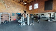 This fitness center masterfully blends historic industrial architecture with modern workout facilities. Housed within a repurposed brick building, the space features soaring ceilings with exposed structural beams and warehouse-style windows that flood the interior with natural light. The original brick walls add striking character throughout, while large glass garage doors and floor-to-ceiling windows along one side offer direct views of an outdoor pool area, seamlessly connecting the interior to the surrounding grounds.

The main workout floor is finished with durable rubber flooring and is generously proportioned to accommodate a wide range of equipment zones. A dedicated strength training area includes cable machines, free weights, adjustable benches, and a full dumbbell rack, complemented by large wall-mounted mirrors that visually expand the space. A cardio section lines one wall with multiple treadmills, ellipticals, and stationary cycling bikes, with wall-mounted televisions positioned for easy viewing. Medicine balls and additional accessories are neatly organized on dedicated racks. An upper mezzanine level, visible from below, overlooks the main floor and adds to the impressive sense of vertical scale. The overall ambiance is energetic yet refined, balancing industrial heritage with contemporary fitness functionality.