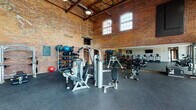This fitness center masterfully blends historic industrial architecture with modern workout facilities. Housed within a repurposed brick building, the space features soaring ceilings with exposed structural beams and warehouse-style windows that flood the interior with natural light. The original brick walls add striking character throughout, while large glass garage doors and floor-to-ceiling windows along one side offer direct views of an outdoor pool area, seamlessly connecting the interior to the surrounding grounds.

The main workout floor is finished with durable rubber flooring and is generously proportioned to accommodate a wide range of equipment zones. A dedicated strength training area includes cable machines, free weights, adjustable benches, and a full dumbbell rack, complemented by large wall-mounted mirrors that visually expand the space. A cardio section lines one wall with multiple treadmills, ellipticals, and stationary cycling bikes, with wall-mounted televisions positioned for easy viewing. Medicine balls and additional accessories are neatly organized on dedicated racks. An upper mezzanine level, visible from below, overlooks the main floor and adds to the impressive sense of vertical scale. The overall ambiance is energetic yet refined, balancing industrial heritage with contemporary fitness functionality.