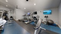 Located at 2025 W Indian School Rd in Phoenix, Arizona, this fitness center offers a well-equipped exercise space designed to support a range of workout routines. The room features durable rubber flooring in a dark gray and blue speckled finish, providing a safe and functional surface for various exercises. Recessed lighting and a ceiling fan keep the space bright and well-ventilated, contributing to a clean, modern aesthetic with crisp white walls and white baseboards throughout.

The cardio section is anchored by multiple treadmills and an elliptical trainer, complemented by a stationary bike and additional cardio equipment. Several wall-mounted flat-screen televisions are positioned throughout the room, allowing users to stay entertained during their workouts. A multi-functional cable crossover machine occupies one end of the room, offering versatile strength training options. A weight bench and additional strength equipment round out the workout offerings.

Large wall mirrors are strategically placed to help users monitor their form and enhance the sense of openness within the space. A glass-paneled entry door allows natural light to filter in and provides a view to the exterior. The overall layout is organized and efficient, making effective use of the available floor area to deliver a comprehensive fitness facility suitable for cardiovascular training, strength building, and general conditioning.