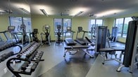 This fitness center offers a comprehensive workout environment designed to accommodate a variety of exercise routines. The space is finished with durable rubber flooring, providing both safety and comfort during workouts. Large windows along the walls allow natural light to fill the room while offering views of the surrounding exterior, creating an open and energizing atmosphere. Ceiling fans are mounted overhead to maintain airflow and comfort throughout the space.

The cardio section is well-stocked with multiple treadmills and elliptical trainers, providing ample options for cardiovascular exercise. A dedicated free weight area lines one wall, featuring an extensive rack of dumbbells in graduated sizes, suitable for a wide range of strength training exercises. Additional strength equipment, including cable machines and adjustable benches, is positioned throughout the room, offering users the tools needed for a full-body workout.

Recessed and fluorescent lighting supplements the natural light, ensuring the space remains well-lit throughout all hours. The layout is thoughtfully organized, providing sufficient space between equipment to allow for comfortable movement and use by multiple individuals simultaneously.