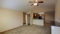 This single-floor space showcases an open-concept layout that seamlessly connects the living area to the kitchen. The living room features vaulted ceilings fitted with a ceiling fan and light fixture, adding a sense of height and airiness to the space. Neutral beige carpeting extends throughout, complementing the warm-toned walls and wood baseboards.

The kitchen is positioned at the rear of the open living area and is defined by a raised breakfast bar with dark countertops, providing a natural division between the two spaces. Warm wood cabinetry lines the kitchen walls, and black appliances add a contrasting accent. Pendant lighting hangs above the counter area, supplemented by a fluorescent fixture overhead for ample task lighting.

A doorway visible from the living area leads to additional rooms, with a closet featuring built-in shelving conveniently located nearby. The overall design prioritizes functionality and an open feel, with the connected layout making the space feel expansive and well-suited for everyday living.