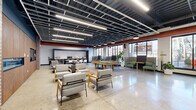 This generously sized single-floor amenity lounge showcases a compelling industrial-modern aesthetic throughout its open-plan layout. Polished concrete floors reflect natural light streaming in through expansive, floor-to-ceiling windows along the exterior wall, offering views of the surrounding streetscape and keeping the space bright and airy.

The exposed ceiling is a defining architectural feature, revealing a dark-painted structural grid, suspended linear LED lighting, and prominent copper-toned HVAC ductwork that adds an industrial character to the overall design. Brick columns punctuate the interior, further reinforcing the building's raw, urban character.

The space is thoughtfully organized into distinct functional zones. A social lounge area features modern upholstered armchairs with wood-frame accents arranged around a central pool table set atop a textured area rug. Semi-private booth seating with high acoustic panels provides quieter nooks along the windowed perimeter, ideal for focused work or small gatherings.

A full kitchen and dining area is equipped with dark cabinetry, appliances, a long communal dining table, and under-counter beverage refrigerators. Warm wood wall paneling and built-in shelving anchor a media lounge zone featuring a wall-mounted television and comfortable sofas, offering a more relaxed retreat within the larger space.