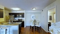 This single-floor residence offers a thoughtfully designed living space that seamlessly blends modern aesthetics with everyday functionality. The kitchen is a standout feature, boasting rich dark cabinetry that contrasts elegantly with granite countertops and stainless steel appliances, including a microwave, range, and refrigerator. A kitchen island provides additional prep space and adds to the open, flowing feel of the layout. The warm hardwood flooring extends throughout, lending a sense of continuity and warmth to the space. Adjacent to the kitchen, the dining area is intimate yet stylish, accommodating a glass-top bistro table paired with upholstered counter-height chairs — ideal for casual meals or entertaining. A decorative sunburst mirror anchors the dining nook, adding a touch of personality to the otherwise clean and neutral palette. Flush-mount ceiling lights in both areas provide consistent, ambient illumination throughout. The walls are finished in crisp white, complementing the textured ceiling and contributing to a bright, airy atmosphere. Overall, the space strikes a balance between contemporary design and comfortable living.