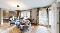 Located at 100 Granby Crossing in Cayce, South Carolina, this thoughtfully designed living room delivers a refined yet functional layout across a single floor. The space is anchored by a striking built-in wood media wall on one side, complete with open shelving, decorative display niches, and integrated lighting beneath the console — providing both a visual focal point and ample storage. Warm, light-toned hardwood flooring runs throughout, lending a sense of cohesion and natural warmth to the room. A statement circular pendant light in a dark bronze finish hangs overhead, adding a contemporary touch to the overall design. On the opposite end, a set of triple windows dressed in floral-patterned drapes and wood-shuttered blinds allows generous natural light to fill the space while maintaining privacy. Beneath the windows sits a rustic shuffleboard table, offering a social and recreational element to the room. A nook along the back wall features a richly textured wood slat accent wall framing a compact workspace with a green chair, adding both texture and function. Plush, upholstered armchairs in muted blue-gray tones are arranged on a vintage-style area rug at the center, creating an inviting seating arrangement ideal for conversation and relaxation.