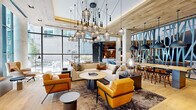 Located at 8005 Gramercy Blvd in Rockville, Maryland, this sophisticated single-floor amenity lounge delivers a refined, hotel-inspired atmosphere through thoughtful design and upscale finishes. The expansive main gathering area is anchored by soaring ceilings and flooded with natural light from floor-to-ceiling windows, offering views of the surrounding exterior. A dramatic cluster of cage-style pendant lights hangs overhead, complementing the warm wood ceiling planks and light herringbone hardwood flooring that lend organic texture throughout the space.

Seating areas are carefully curated with plush sectional sofas, bold amber-hued lounge chairs, and layered geometric-patterned rugs, creating inviting pockets for relaxation or socializing. Decorative steel partition screens with abstract botanical motifs elegantly divide zones while maintaining an open, airy feel. Built-in shelving units framed in matte black steel display curated décor objects and books, surrounding a wall-mounted flat-screen television in a cozy media lounge nook.

A high-top communal table with bar seating provides a casual gathering spot, while an adjoining game room features a full-size billiards table, additional wall-mounted displays, and stylish pendant lighting. The cohesive design palette of warm amber, natural wood, concrete, and dark steel accents creates a dynamic yet welcoming environment suited for both leisure and community engagement.