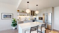 This thoughtfully designed one-bedroom, one-bathroom residence offers a cohesive open-concept layout that maximizes every square foot of its efficient footprint. The combined kitchen and living area serves as the heart of the home, anchored by a sleek waterfall-edge island with bar seating beneath a pair of globe pendant lights. The kitchen features light wood cabinetry, a textured white hexagonal tile backsplash, quartz countertops, stainless steel appliances including a range with hood and built-in microwave, and open floating shelves for display. The living area extends naturally from the kitchen and is defined by large floor-to-ceiling windows framed in matte black, flooding the space with natural light and offering dramatic views of the surrounding mountain range and city skyline. Hardwood-style flooring runs throughout the main living areas, lending warmth and continuity. The bedroom is a calm, neutral retreat with plush carpet, a large window capturing the same elevated views, and a dedicated closet for ample storage. The bathroom is finished with tile flooring, a wood-toned vanity with a white countertop, and a full tub-shower combination with large-format tile surround.