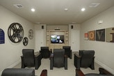 This single-level media center is designed as a private screening room with a clear cinematic theme throughout. The room is outfitted with rows of dark navy blue theater-style recliners, each equipped with built-in cup holders and armrests, providing a comfortable and immersive viewing experience for multiple guests. The seating is arranged in a forward-facing layout directed toward a built-in wooden entertainment center housing a large flat-screen television, flanked by storage cabinetry below and open shelving above.

The room's décor reinforces its movie-centric identity, featuring oversized vintage film reels mounted on the walls alongside colorful typographic canvas art panels spelling out "MOVIES." Surround sound speakers are strategically positioned around the room to enhance the audio experience. Recessed lighting in the smooth ceiling provides ambient illumination, while neutral beige walls and carpeted flooring create a warm, theater-like atmosphere.

A paneled white door connects the screening room to an adjacent common area, which features stone column accents and additional amenities, suggesting this media center is part of a larger shared facility. The overall design balances comfort and functionality, making it well-suited for group screenings and entertainment.