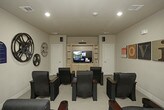 This single-level media center is designed as a private screening room with a clear cinematic theme throughout. The room is outfitted with rows of dark navy blue theater-style recliners, each equipped with built-in cup holders and armrests, providing a comfortable and immersive viewing experience for multiple guests. The seating is arranged in a forward-facing layout directed toward a built-in wooden entertainment center housing a large flat-screen television, flanked by storage cabinetry below and open shelving above.

The room's décor reinforces its movie-centric identity, featuring oversized vintage film reels mounted on the walls alongside colorful typographic canvas art panels spelling out "MOVIES." Surround sound speakers are strategically positioned around the room to enhance the audio experience. Recessed lighting in the smooth ceiling provides ambient illumination, while neutral beige walls and carpeted flooring create a warm, theater-like atmosphere.

A paneled white door connects the screening room to an adjacent common area, which features stone column accents and additional amenities, suggesting this media center is part of a larger shared facility. The overall design balances comfort and functionality, making it well-suited for group screenings and entertainment.