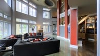This striking contemporary lounge space makes an immediate impression with its dramatic double-height ceilings and an abundance of natural light streaming through expansive floor-to-ceiling windows. The architectural design is bold and intentional, showcasing a palette of cool gray walls offset by vivid red accent columns that draw the eye upward and create strong visual anchors throughout the space. A decorative circular ceiling medallion adds a refined architectural detail overhead, complementing the modern aesthetic. The polished concrete flooring reflects light beautifully, enhancing the open, airy atmosphere of the room. Seating is arranged thoughtfully with dark charcoal sofas accented by red, black, and white throw pillows, creating a cohesive and stylish lounge environment. Sculptural artwork and carefully placed accent lighting contribute to the space's artistic character. A mezzanine-level overlook is visible above, adding depth and verticality to the overall layout. A flat-screen display is integrated into the feature wall near the columns, suggesting a multifunctional use for gatherings or presentations. Large windows frame views of lush greenery outside, connecting the interior to the natural surroundings and reinforcing the bright, welcoming ambiance of this well-appointed communal space.