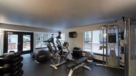 This dedicated fitness space is designed to support a well-rounded workout routine. The room is outfitted with a selection of cardio equipment, including an elliptical trainer, a stationary cycle, and a treadmill, arranged to make efficient use of the available floor space. A multi-function cable and weight machine anchors one side of the room, complemented by a workout bench, a stability ball, and additional accessories for strength and flexibility training. A wall-mounted flat-screen television provides entertainment during workouts. The space benefits from generous natural light, with large windows lining multiple walls and offering views of the surrounding outdoor environment, including lush greenery visible through one set of windows. The light-toned walls and smooth ceiling contribute to a clean, open atmosphere, while the dark rubberized flooring provides a practical, durable surface suited for exercise. The overall design is functional and purposeful, creating an environment well-suited for daily fitness activity.