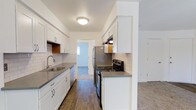 This single-floor space offers a clean, modern aesthetic throughout its well-designed layout. The kitchen serves as a standout feature, showcasing white shaker-style cabinetry that extends to the upper and lower walls, providing generous storage. Grey laminate countertops offer ample prep space, while a classic white subway tile backsplash adds a timeless, polished touch. Stainless steel appliances, including a refrigerator, electric range, and dishwasher, complement the contemporary design. A stainless steel sink with a modern faucet is positioned beneath a window, allowing natural light to filter into the space. The kitchen flows seamlessly into adjacent rooms, with hardwood-style flooring underfoot that transitions into carpeted areas visible through the open doorway. The overall color palette is neutral and bright, with white walls and ceilings that enhance the sense of openness. The layout is functional and efficient, with a corridor-style kitchen that connects to the rest of the living areas. The space presents a fresh, move-in-ready environment that balances practicality with modern design sensibilities.